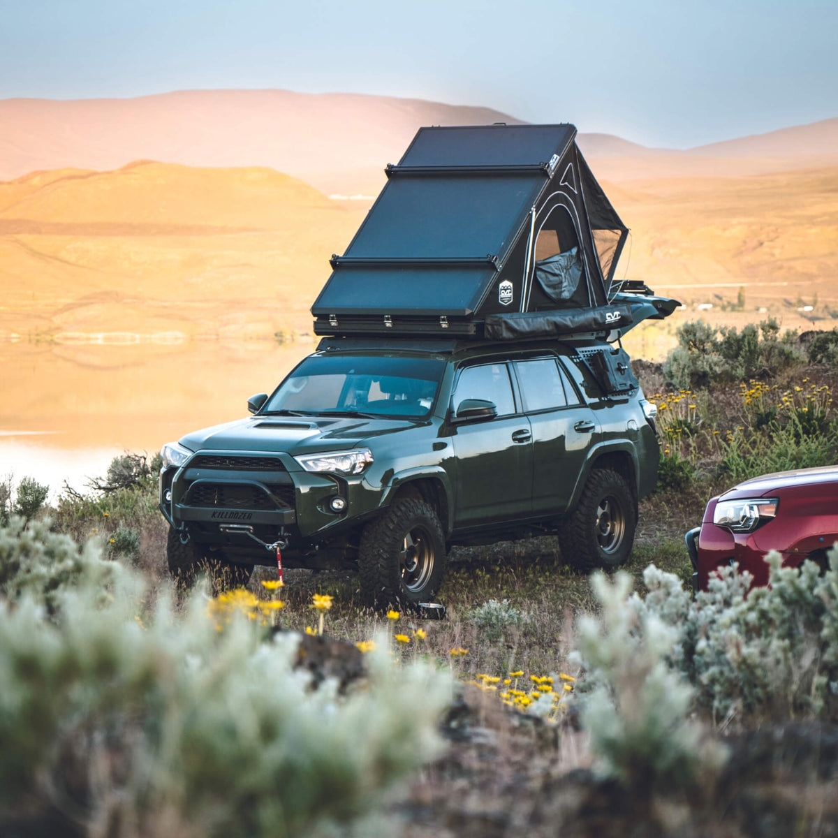 A dark green SUV with a rooftop tent is parked outdoors among wildflowers and shrubs, with mountains and a lake in the background at sunset.