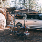 A silver truck equipped with a rooftop tent and the CVT Pioneer Awning - 79″ Medium is parked at a campsite. Beside the truck, a folding table arranged with cooking gear, cups, and food items sits under tall trees near large rocks, providing a comfortable outdoor shelter for relaxing and dining.