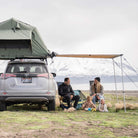 A family with two young children and a dog sits beside their car, which is equipped with a rooftop tent and a CVT Pioneer Awning - 79″ Medium, as they camp by a lake with snow-capped mountains in the background on a cloudy day.