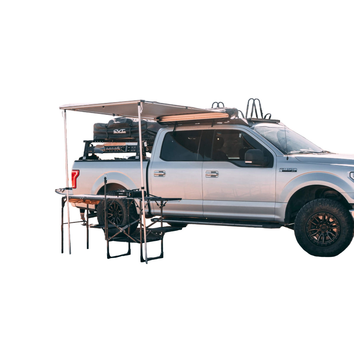 A silver pickup truck with black wheels is parked with a CVT Pioneer Awning - 79″ Medium extended from its side, creating a shaded outdoor setup with a table and chairs underneath. Camping gear is stored on the truck bed and roof rack.