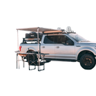 A silver pickup truck with black wheels is parked with a CVT Pioneer Awning - 79″ Medium extended from its side, creating a shaded outdoor setup with a table and chairs underneath. Camping gear is stored on the truck bed and roof rack.