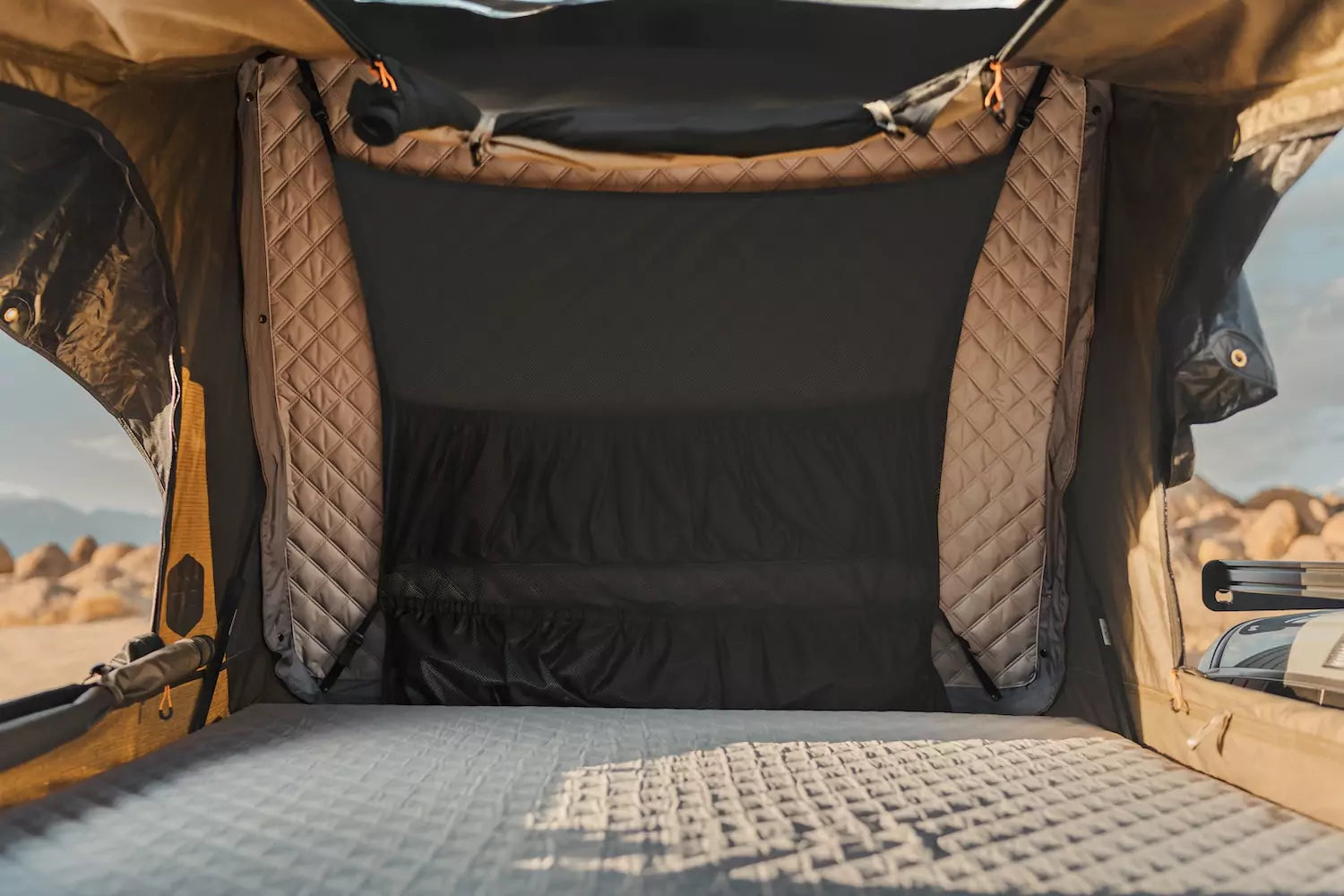 Interior view of a rooftop tent with quilted walls and mattress, set up outdoors with a rocky landscape visible through the open sides.