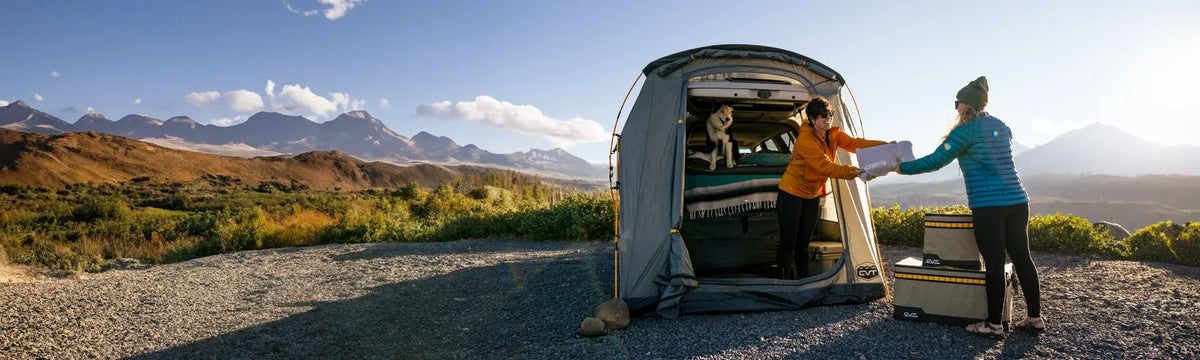 Two people set up camp beside a large tent with a dog inside, surrounded by mountains, greenery, and a bright sky. One person hands an item to the other near camping gear.
