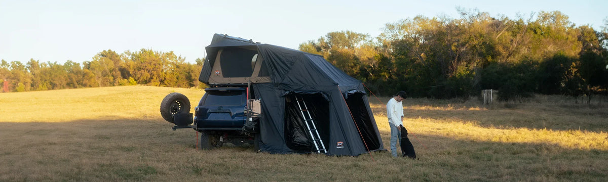 A person stands near a black SUV with a rooftop tent and awning setup in a grassy field, surrounded by trees under a clear sky.