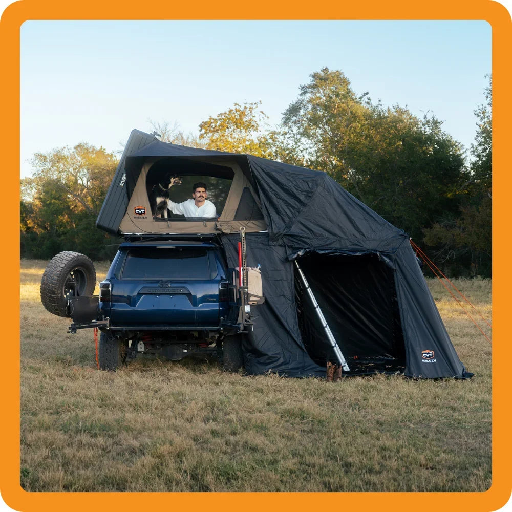 A person waves from a rooftop tent mounted on a dark SUV with an attached awning, parked on dry grass in a field with green trees in the background. The image has an orange border.