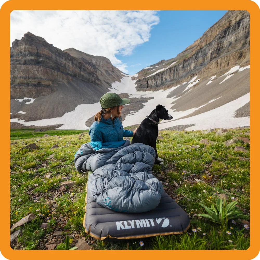 A person in outdoor gear sits on a sleeping pad with a dog, surrounded by grass, mountains, and patches of snow under a partly cloudy sky.