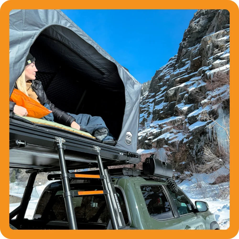 A person sits in a rooftop tent on a green vehicle, looking out at a snowy mountain landscape with rock cliffs and icicles under a bright blue sky.