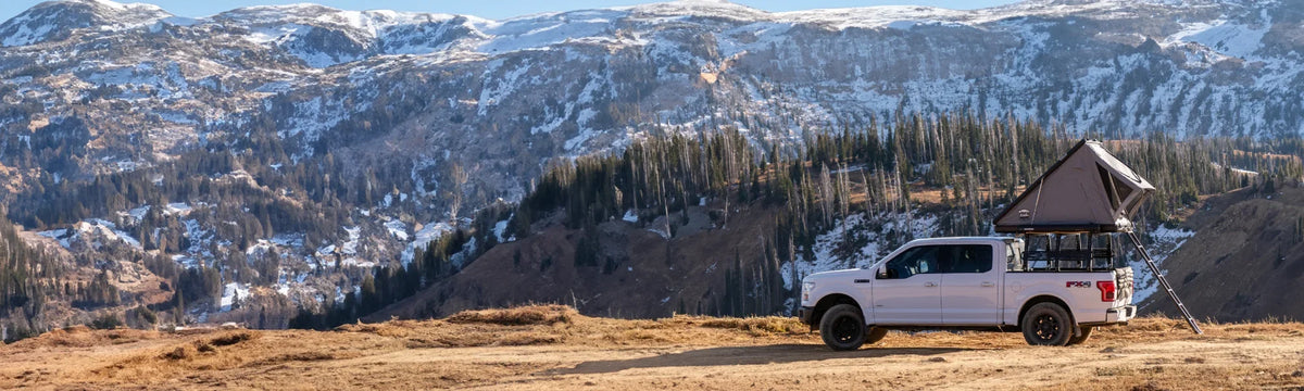 A white pickup truck with a rooftop tent is parked on a dirt road overlooking a scenic mountain landscape with snow-capped peaks and evergreen trees.
