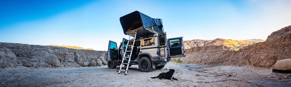 A van with a rooftop tent is parked in a rocky desert area. The vans doors are open, a ladder leads up to the tent, and a black dog is lying on the ground nearby under a clear blue sky.