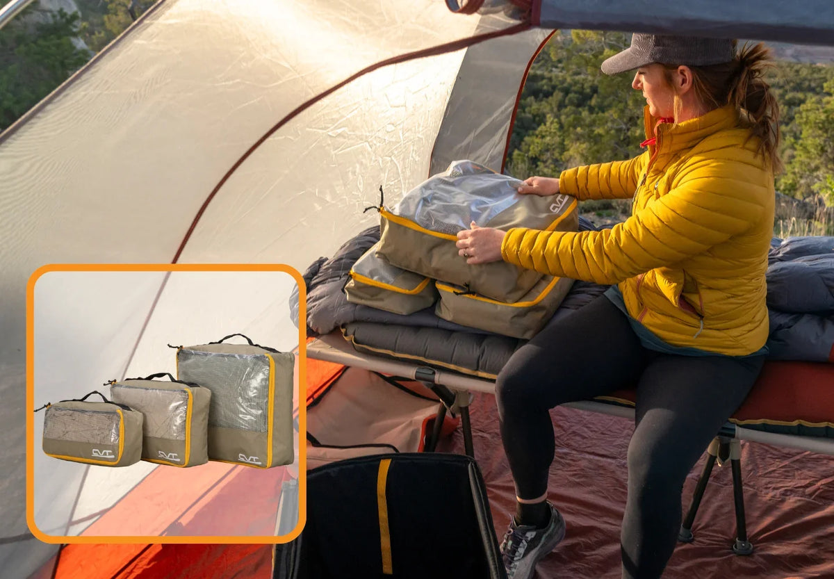 A person in a yellow jacket organizes gray and yellow packing cubes inside a tent. An inset shows close-ups of three stacked packing cubes with zippers and handles. Trees are visible outside the tent.