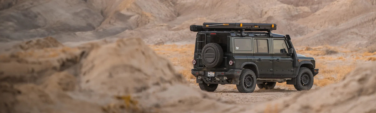A black off-road SUV with a roof rack is parked on rocky terrain in a desert landscape with sandy hills in the background.