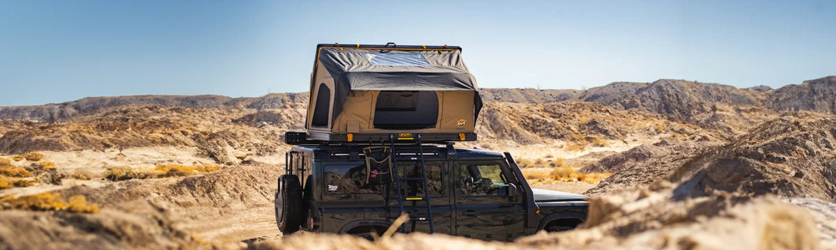 A black SUV with a rooftop tent is parked in a dry, rocky desert landscape under a clear blue sky, surrounded by barren hills and sandy terrain.