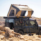 A black off-road vehicle is parked on rocky desert terrain under a clear sky, equipped with the CVT La Sal Hardshell Tent featuring a side-open design and ladder, providing spacious comfort for adventurous travelers.