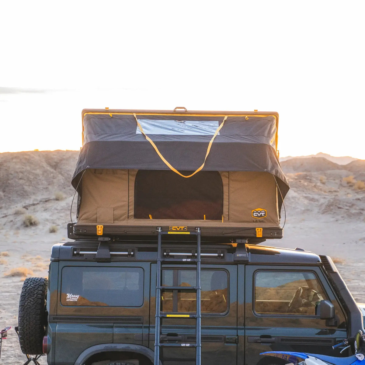 A CVT La Sal Hardshell Tent with a convenient side-open design is set up on top of a black off-road vehicle parked in a desert landscape, with a ladder leading up to the open tent. The sun is setting in the background, highlighting the spacious comfort awaiting adventurers after a long day of exploration.