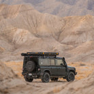A CVT La Sal Hardshell Tent is mounted on the roof rack of a rugged off-road SUV parked on a sandy trail in a barren, mountainous desert landscape beneath an overcast sky.