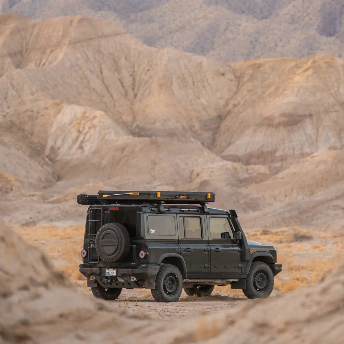 A CVT La Sal Hardshell Tent is mounted on the roof rack of a rugged off-road SUV parked on a sandy trail in a barren, mountainous desert landscape beneath an overcast sky.