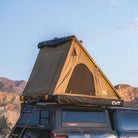 The CVT Mt. Hood 2 Hardshell Tent is mounted on top of a parked vehicle in a desert landscape, ideal for outdoor adventures, with a ladder providing easy access to the tent. Mountains and a clear blue sky form the stunning backdrop.