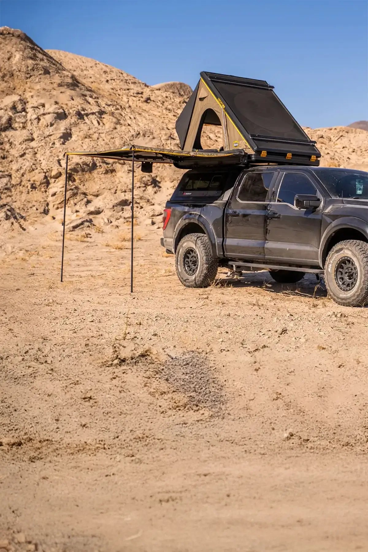 A black pickup truck with a rooftop tent and awning is parked in a desert landscape with rocky hills in the background under a clear sky.
