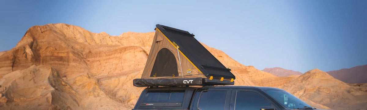 A black truck with a rooftop tent parked in a rocky desert landscape with tan mountains under a clear blue sky.