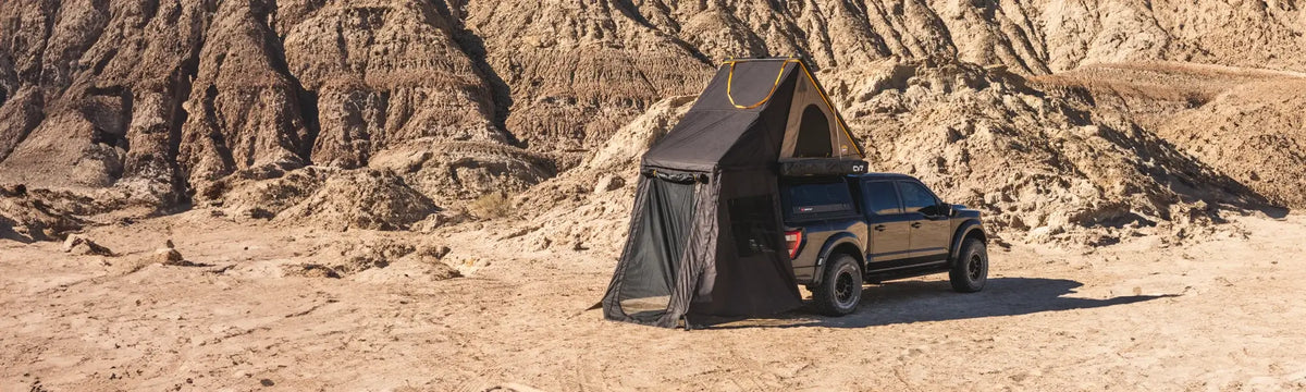 A black pickup truck with a rooftop tent and mesh annex is parked on sandy ground in a rocky desert landscape with rugged hills in the background.