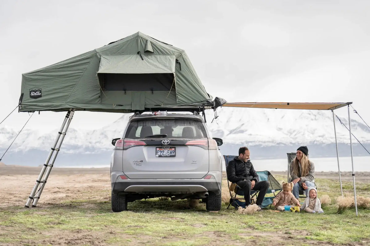 A family of four with a dog sits under an awning attached to an SUV with a rooftop tent, outdoors near mountains and a lake, enjoying a camping trip.