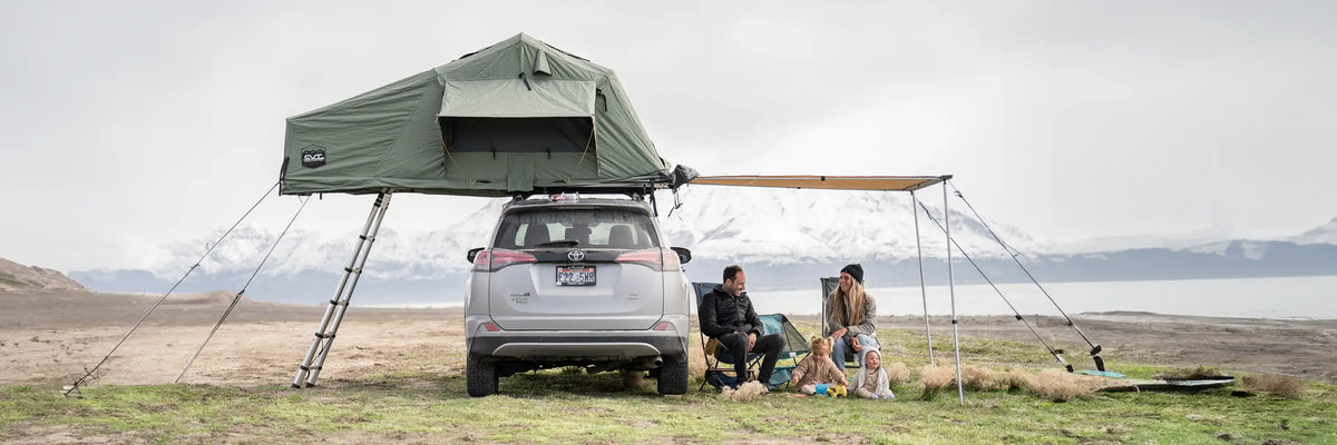 Two people sit by a car with a rooftop tent and awning, camping near a lake with mountains in the background under a cloudy sky. Camping gear is scattered around them.