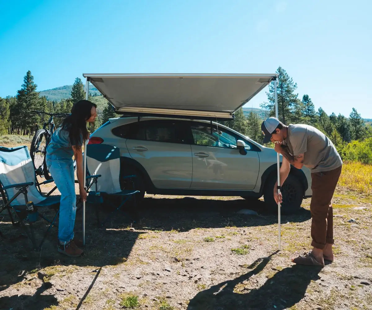 Two people set up a canopy attached to a light-colored car in a sunny outdoor area with trees and mountains in the background. Camping chairs and a bicycle are nearby.