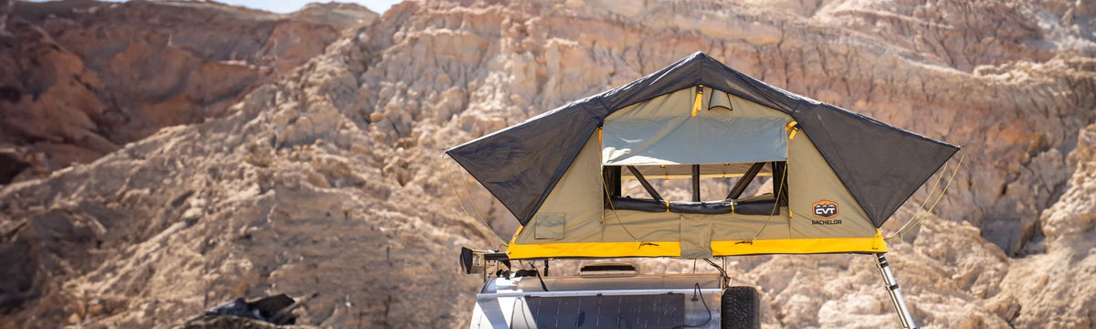A yellow and gray rooftop tent is set up on top of a vehicle, with rugged, rocky desert mountains in the background under bright sunlight.