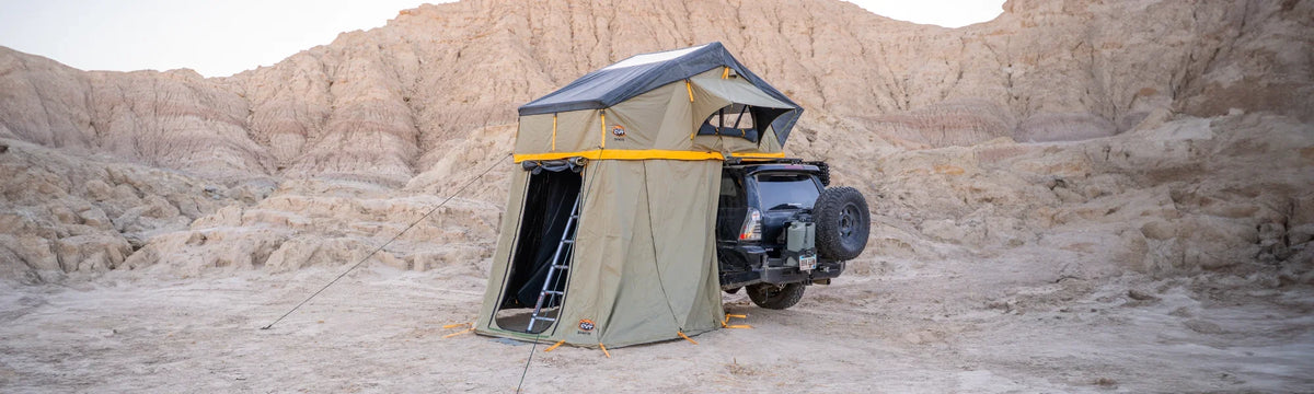 A rooftop tent with an extended annex is set up on an off-road vehicle in a rocky desert landscape, with mountains in the background.