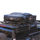 A black SUV with a black rooftop cargo bag secured on a roof rack, featuring the CVT Pioneer Travel Cover Strap Set from the Pioneer tent accessories collection for added security. Traction boards are mounted on the vehicle’s side. The background is plain white.
