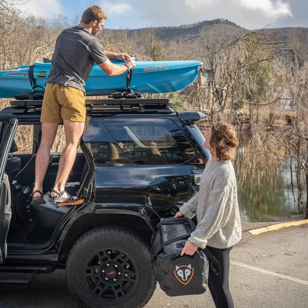 A man stands beside a black SUV, using the Rightline Gear® Moki® Ascent by Rightline Gear to easily secure a blue kayak to the roof rack. Nearby, a woman holding a large black bag looks on as trees and a lake in the background showcase how the Moki® Ascent provides convenient roof access for outdoor adventures.