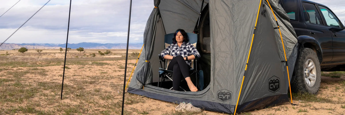 A person sits in a camping chair inside a tent attached to an SUV in a dry, open landscape with mountains in the background.