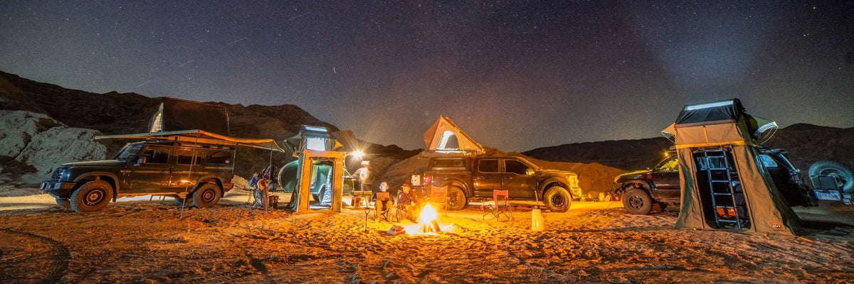 Three off-road vehicles with rooftop tents are parked in a desert at night. Several people are sitting and standing around a campfire under a starry sky, with gear and chairs scattered nearby. Rocky hills are in the background.