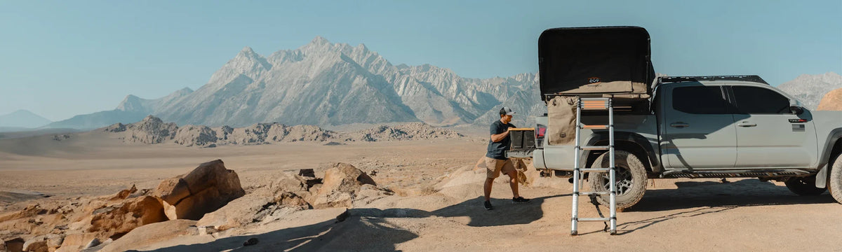A person stands near a gray pickup truck with a rooftop tent set up, surrounded by desert landscape and distant rugged mountains under a clear blue sky.