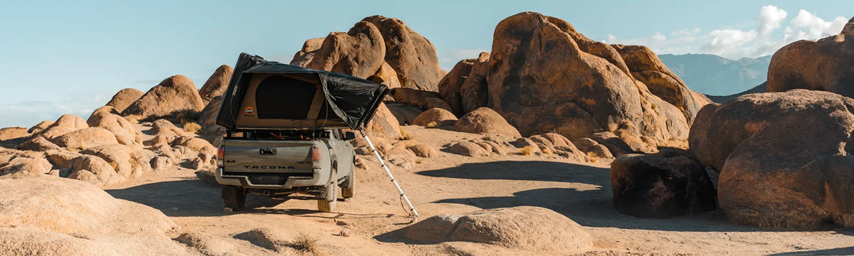 A pickup truck with a rooftop tent is parked among large, sunlit desert boulders under a clear blue sky.
