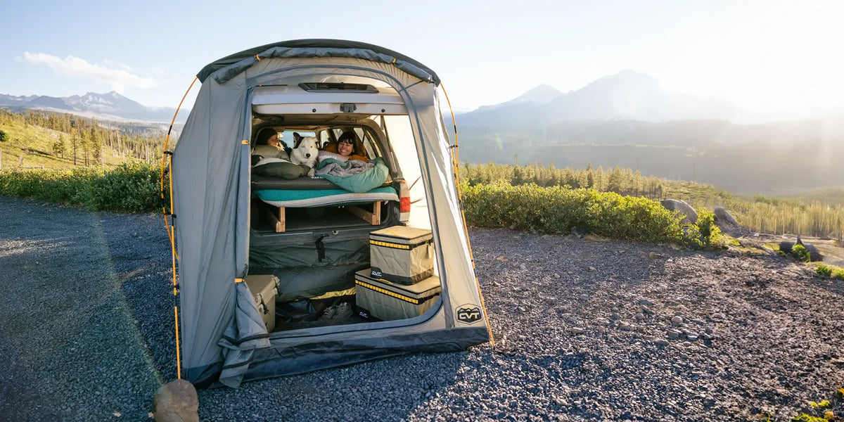 A person and a white dog relax in a rooftop tent on a vehicle, surrounded by camping gear, with mountains and greenery in the background under a clear, sunny sky.