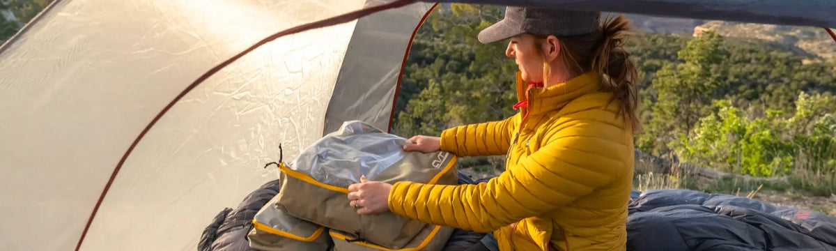 A person in a yellow jacket sits inside a tent, organizing gear in a bag, with trees and hills visible outside in the background.
