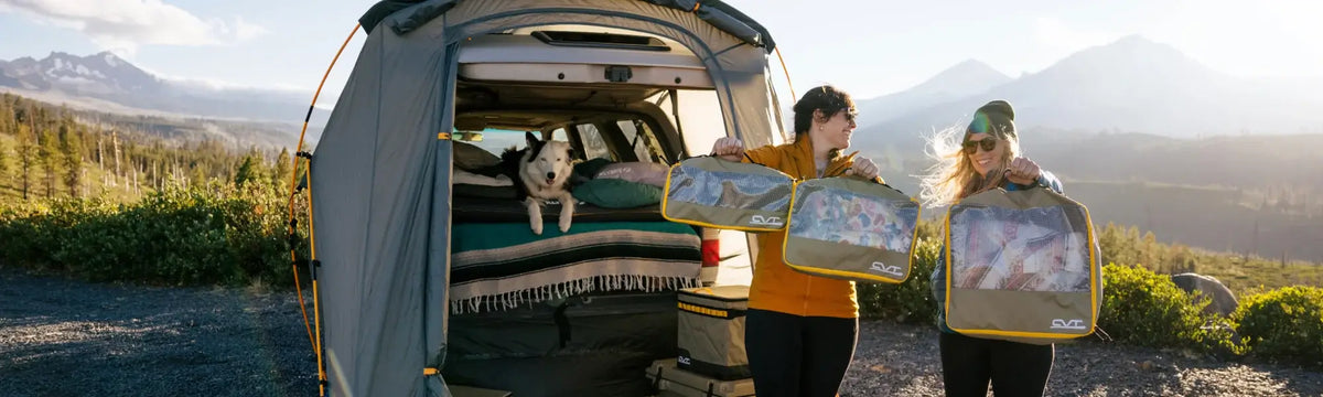 Two people stand by an open tent attached to a vehicle, smiling and holding packing cubes. A dog sits inside the tent. Mountains and greenery are visible in the background under a clear sky.