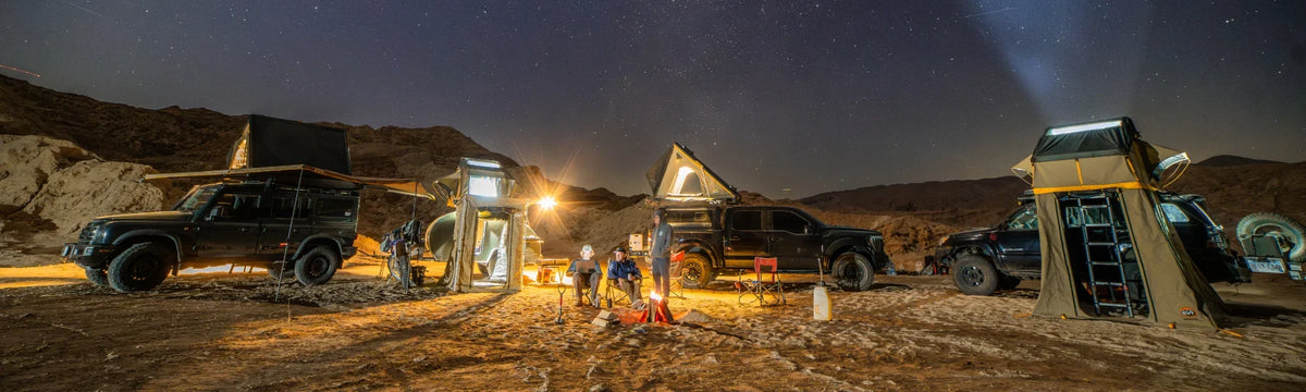 Three off-road vehicles with rooftop tents are parked in a desert at night. People gather around a campfire. Mountains and a starry sky are in the background. Camping gear and lights create a cozy atmosphere.