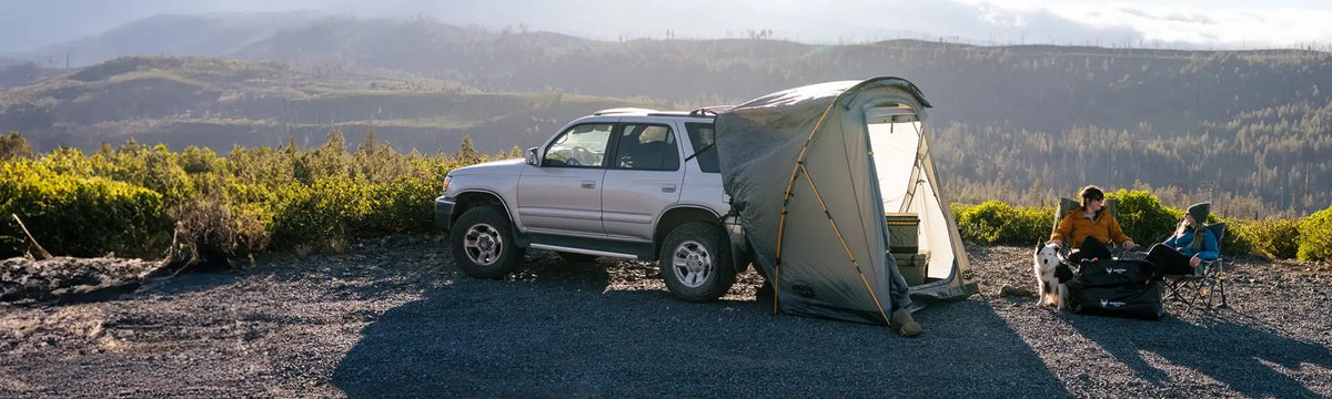 A silver SUV with an attached tent is parked on gravel. Two people and a dog sit nearby on camping chairs, overlooking green hills and mountains in the background under a clear sky.