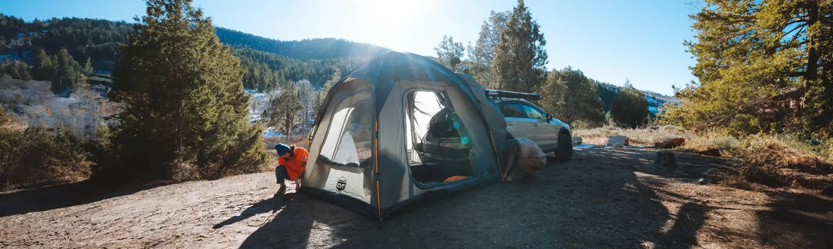 Two people set up a large tent at a sunny, forested campsite near a white car, with mountains and pine trees in the background.