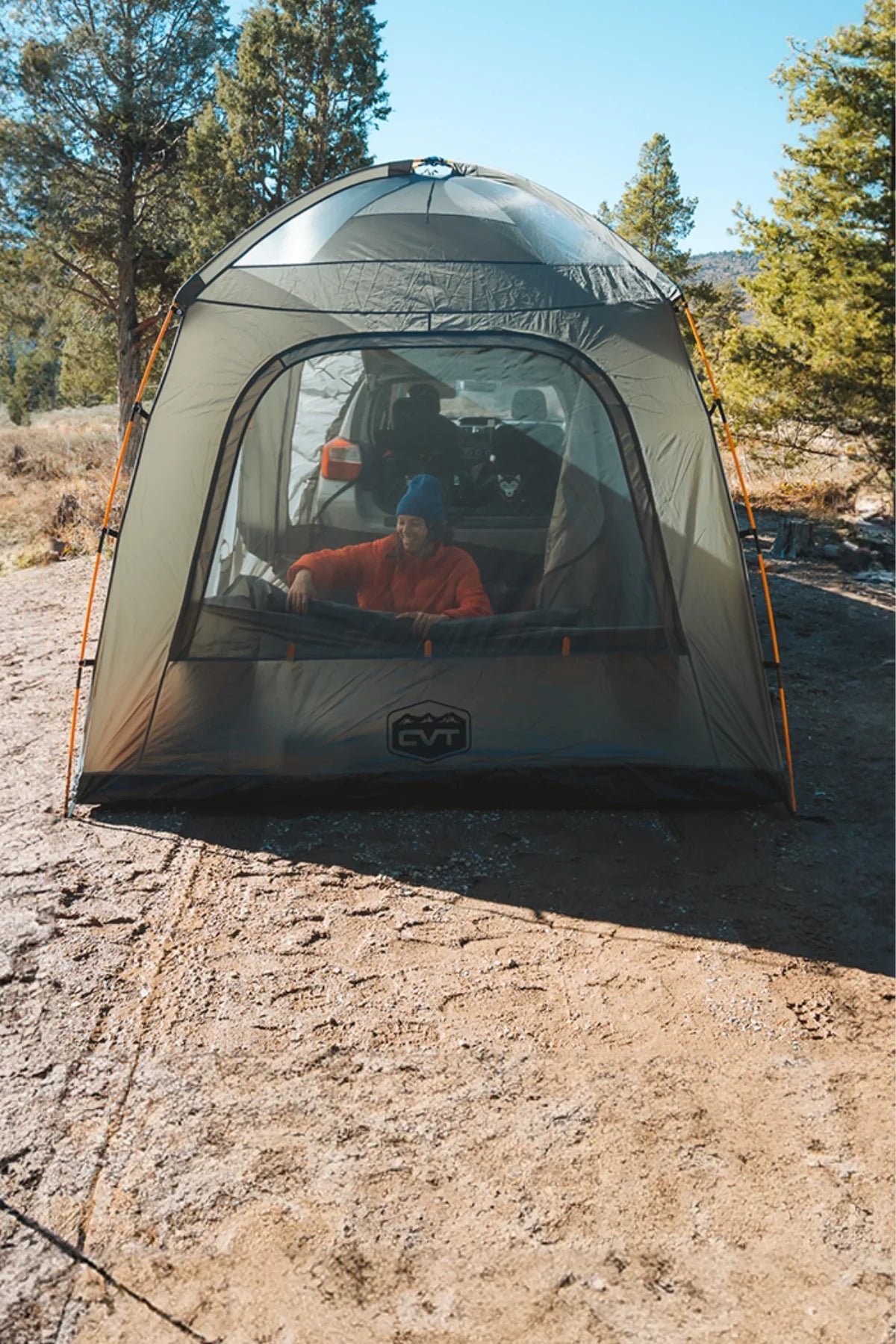 A person wearing a blue beanie sits inside a transparent tent pitched outdoors on a sunny day, surrounded by trees and rocky ground.