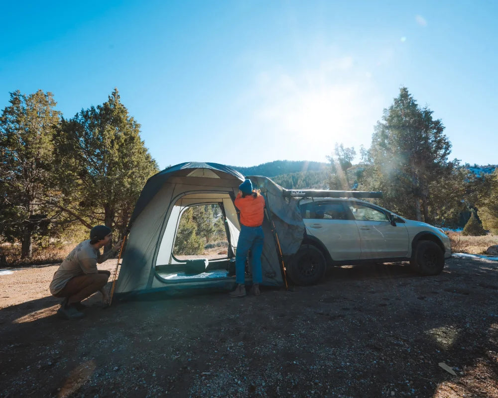 Two people set up a gray tent attached to a white car in a sunny, wooded outdoor area, with trees and hills in the background.