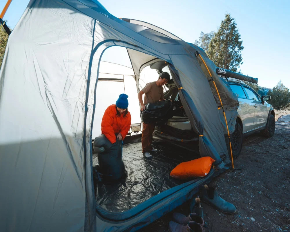 Two people pack gear inside a tent attached to a car. One is kneeling with a black bag, the other is standing, both inside the spacious tent on a dirt campsite surrounded by trees and outdoor gear.