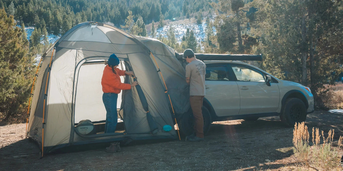 Two people attach a gray tent to the side of a silver car at a forested campsite. Snow patches are visible on the ground and trees in the background under clear daylight.