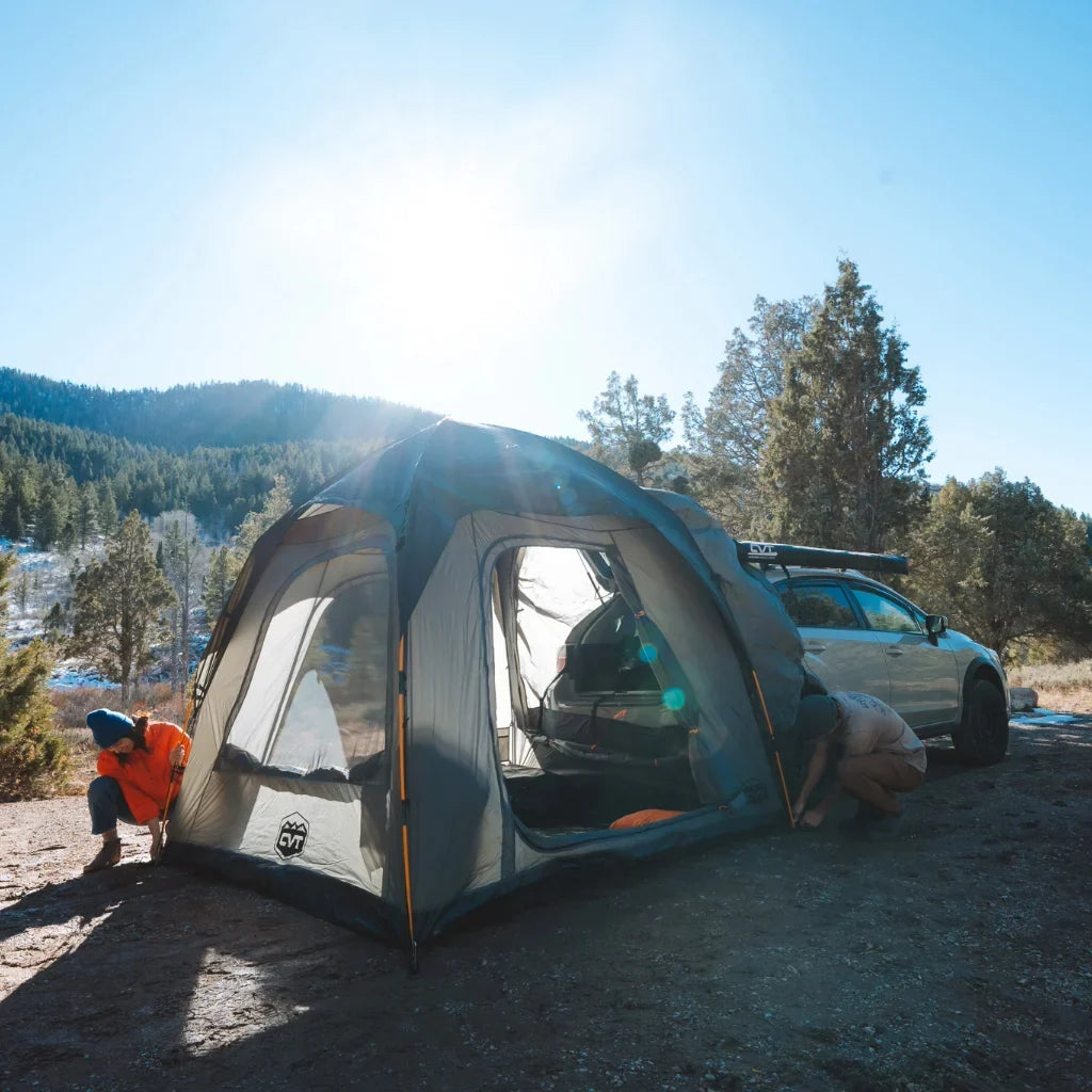 A person in an orange jacket sets up a tent next to a white car in a sunny, forested mountain area. Camping gear is visible; the sky is clear and blue.