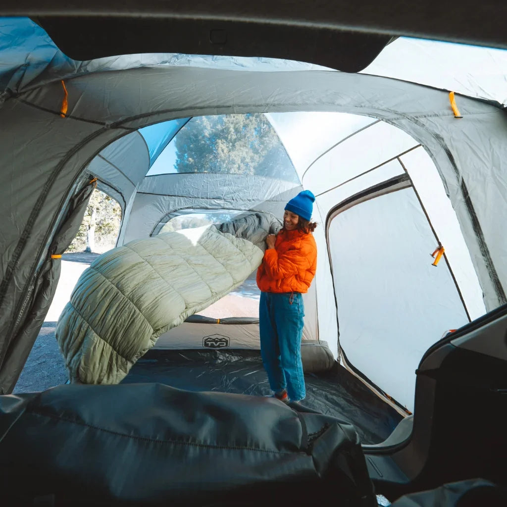 A person in a blue hat and orange jacket sets up a sleeping bag inside a large, light-filled camping tent, with outdoor scenery visible through the windows.