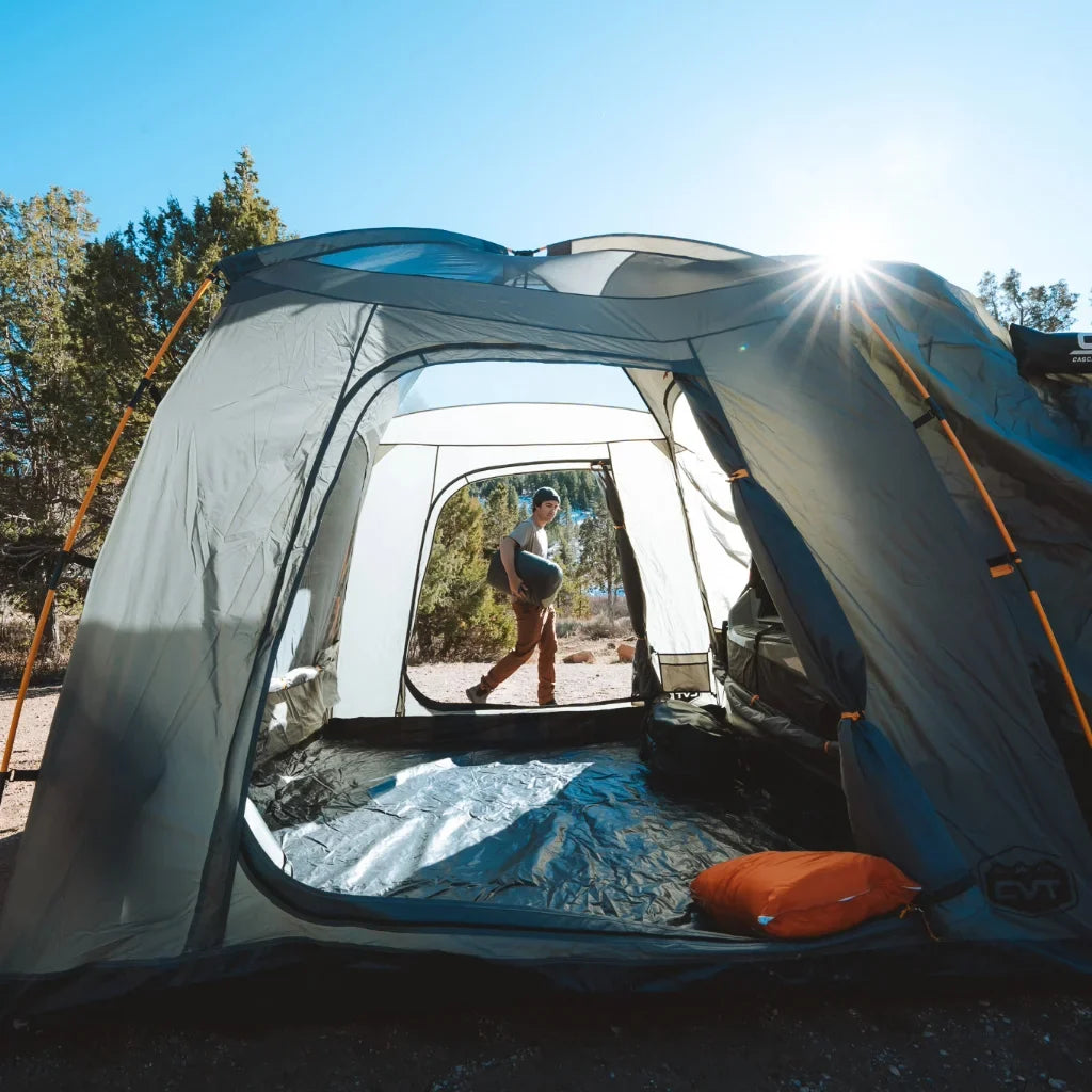 View from inside a large tent looking out at a person standing outside in nature, with trees and bright sunlight in the background. Camping gear is scattered on the tent floor.