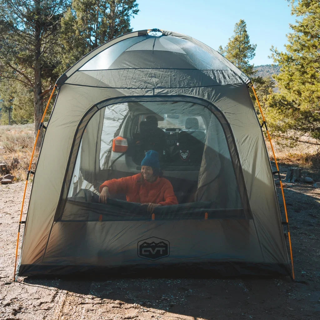 A person in a red jacket and blue beanie sits inside a large, see-through camping tent set up outdoors among trees, with camping gear visible behind them on a sunny day.
