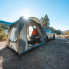 A person wearing an orange jacket relaxes inside the CVT Timber Creek SUV Tent, which is attached to a car at a picturesque outdoor campsite filled with trees and mountain views—a perfect setup for overlanders looking for both adventure and comfort.